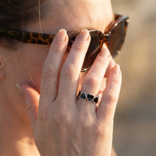 Person wearing sunglasses with a hand holding them, wearing a tarnish-free ring with heart designs.
