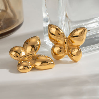 Gold butterfly-shaped earrings on a white surface with a glass container in the background.