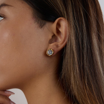 Close-up of a woman wearing gold earrings with a clear gemstone on a neutral background