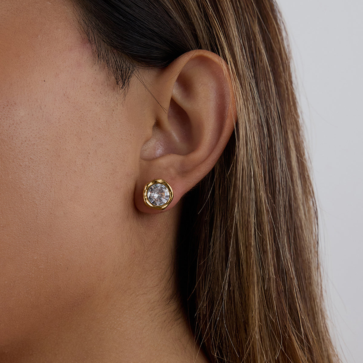 Close-up of an ear wearing a gold earring with a clear gemstone on a neutral background