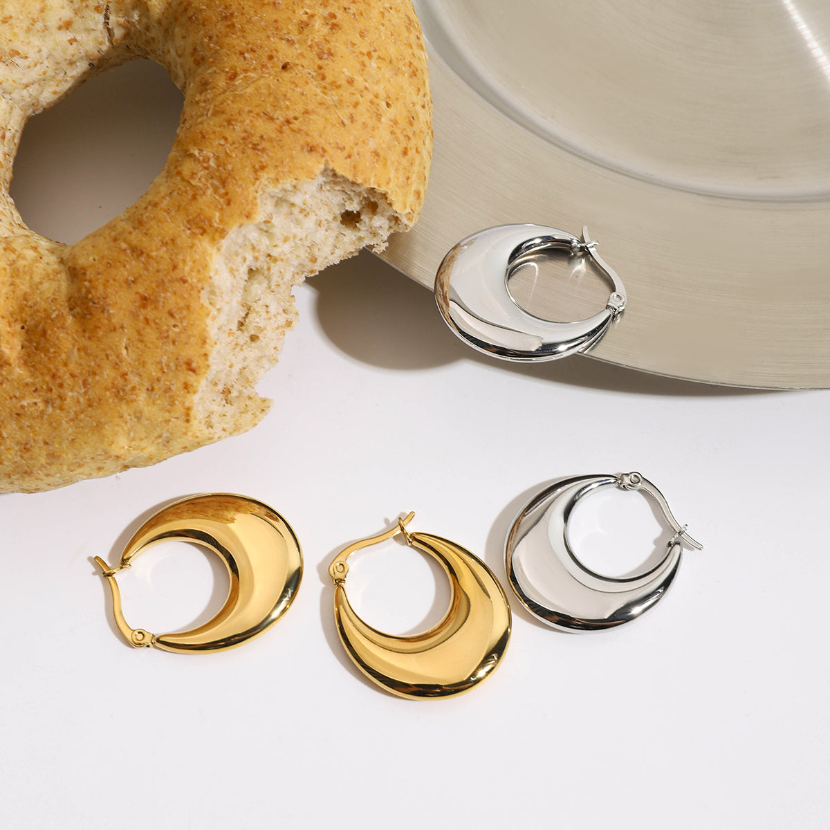 Two pairs of hoop earrings in gold, silver, and bronze on a white surface with a bagel and bowl in the background.