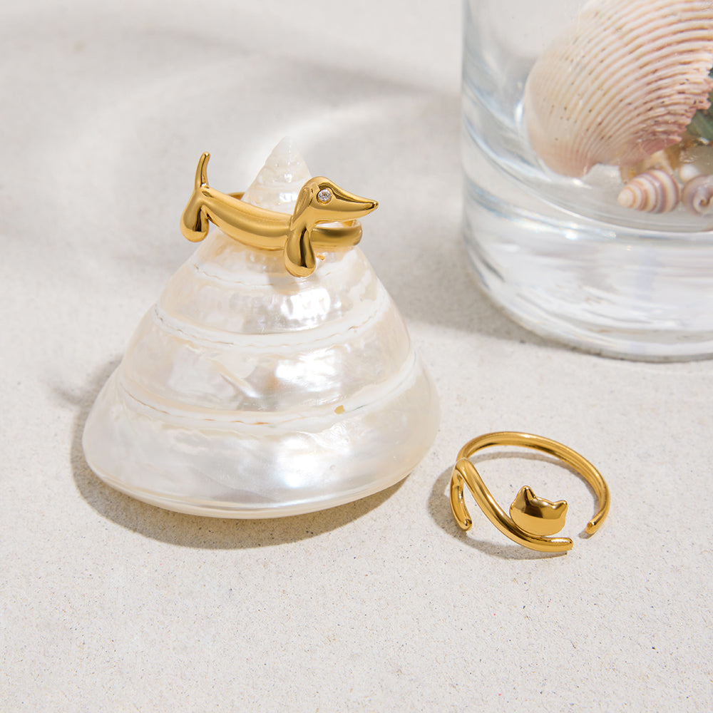 Gold ring shaped like a dachshund on a shell with a glass container, gold ring shaped like a cat and shell in the background.