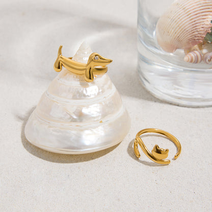 Gold ring shaped like a dachshund on a shell with a glass container, gold ring shaped like a cat and shell in the background.