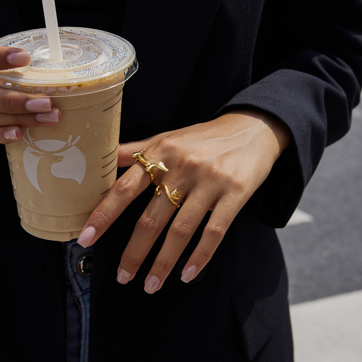 Person holding a Starbucks iced coffee with a straw, wearing gold rings in the shape of cat and dachshund dog