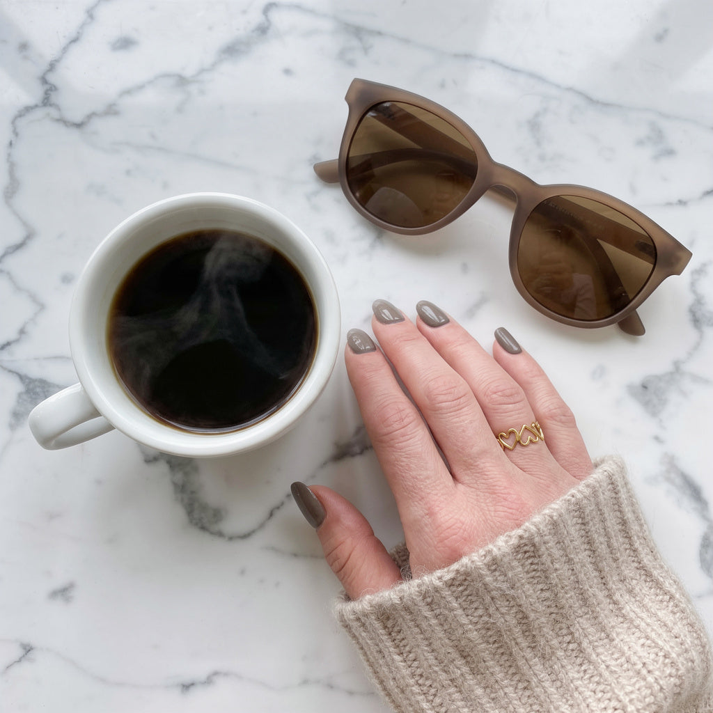 White mug with coffee, brown sunglasses, and hand with beige sleeve and a heart-shaped ring on marble surface