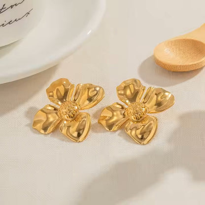 Gold stainless steel flower-shaped earrings on a light surface with a spoon and cup in the background.