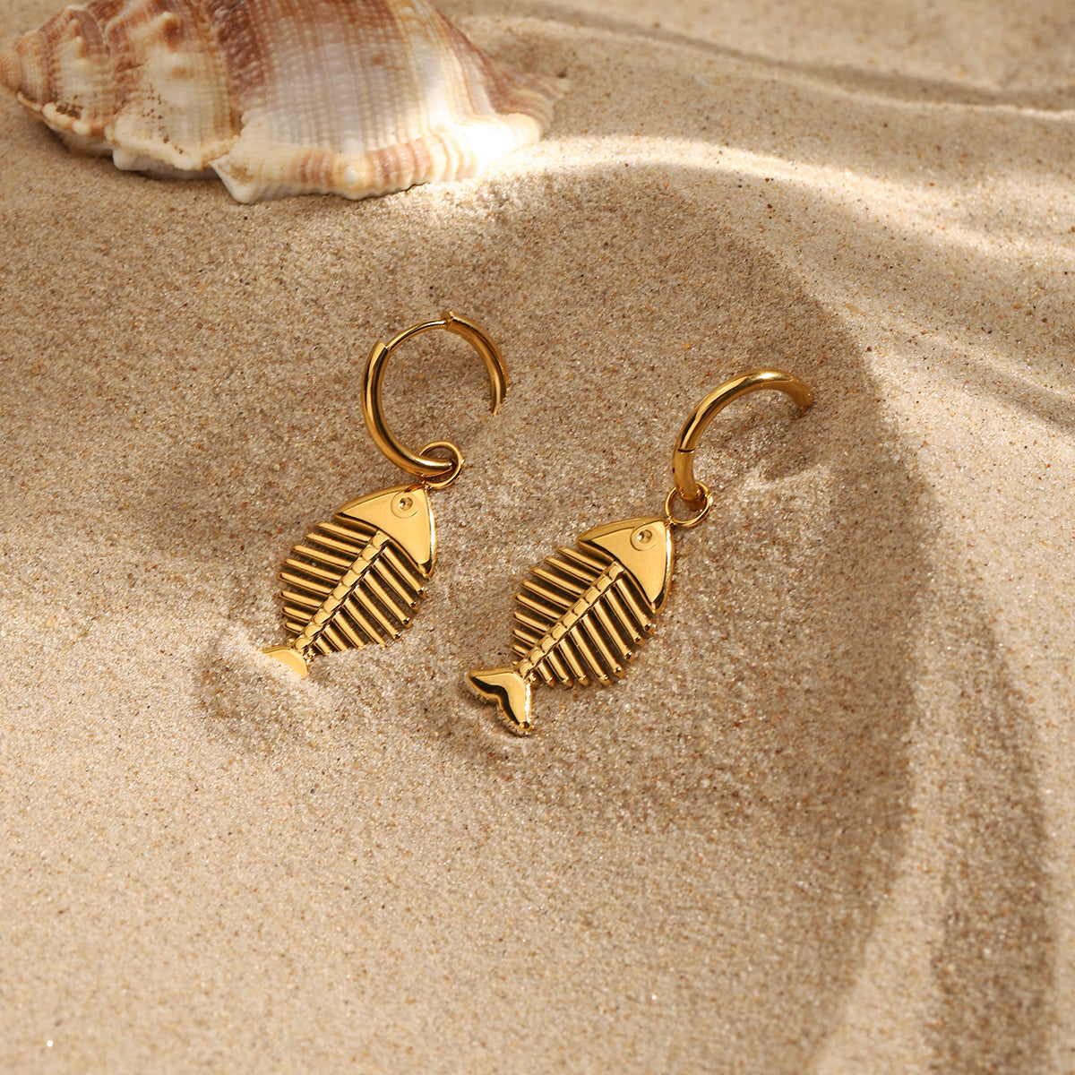 Gold fish-shaped earrings on sand with a shell in the background