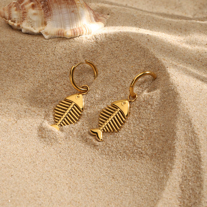 Gold fish-shaped earrings on sand with a shell in the background