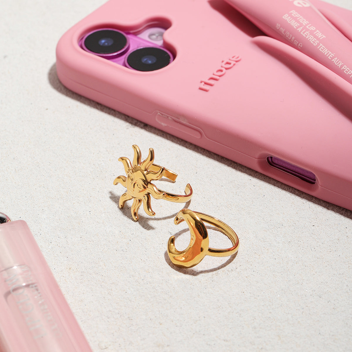 Gold sun and moon ring on a white surface with a pink phone case in the background.