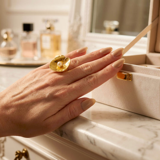 Hand wearing a gold statement ring with a pearl, holding a small box on a marble surface with perfume bottles in the background.