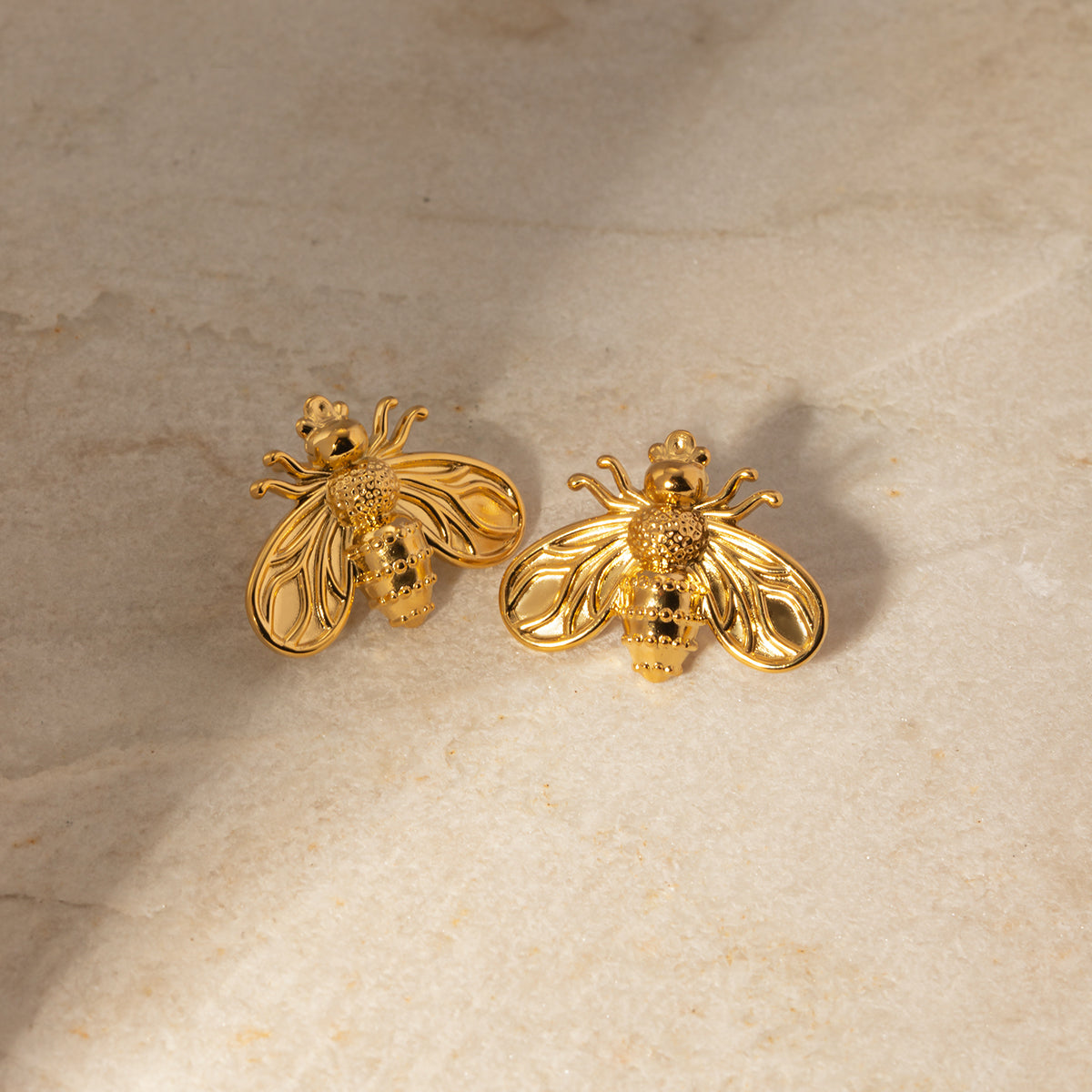 Gold bee-shaped earrings on a beige stone surface
