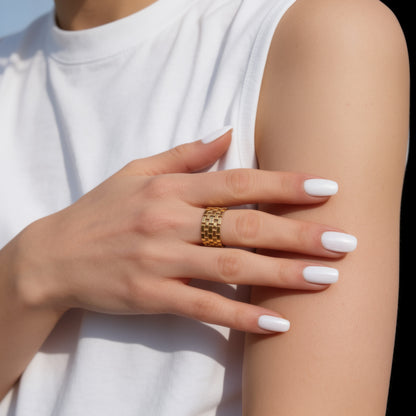 Close-up of a hand wearing a gold ring with white nail polish on a neutral background