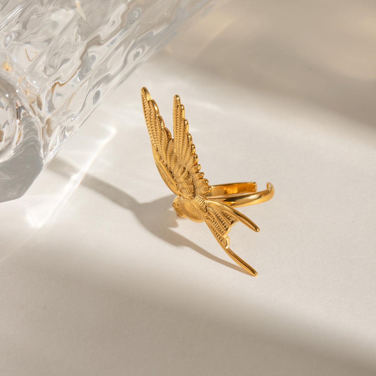 Gold bird-shaped ring on a reflective surface with a glass object in the background