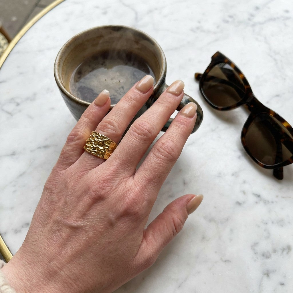 Hand with gold statement ring on marble surface with coffee mug and sunglasses
