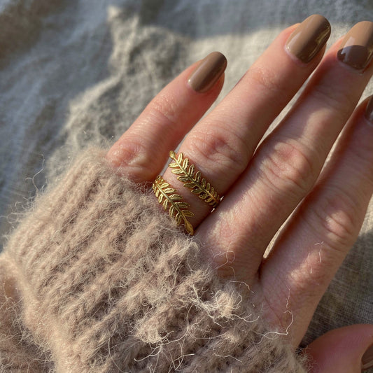 Hand wearing a gold leaf ring with brown nail polish against a textured background