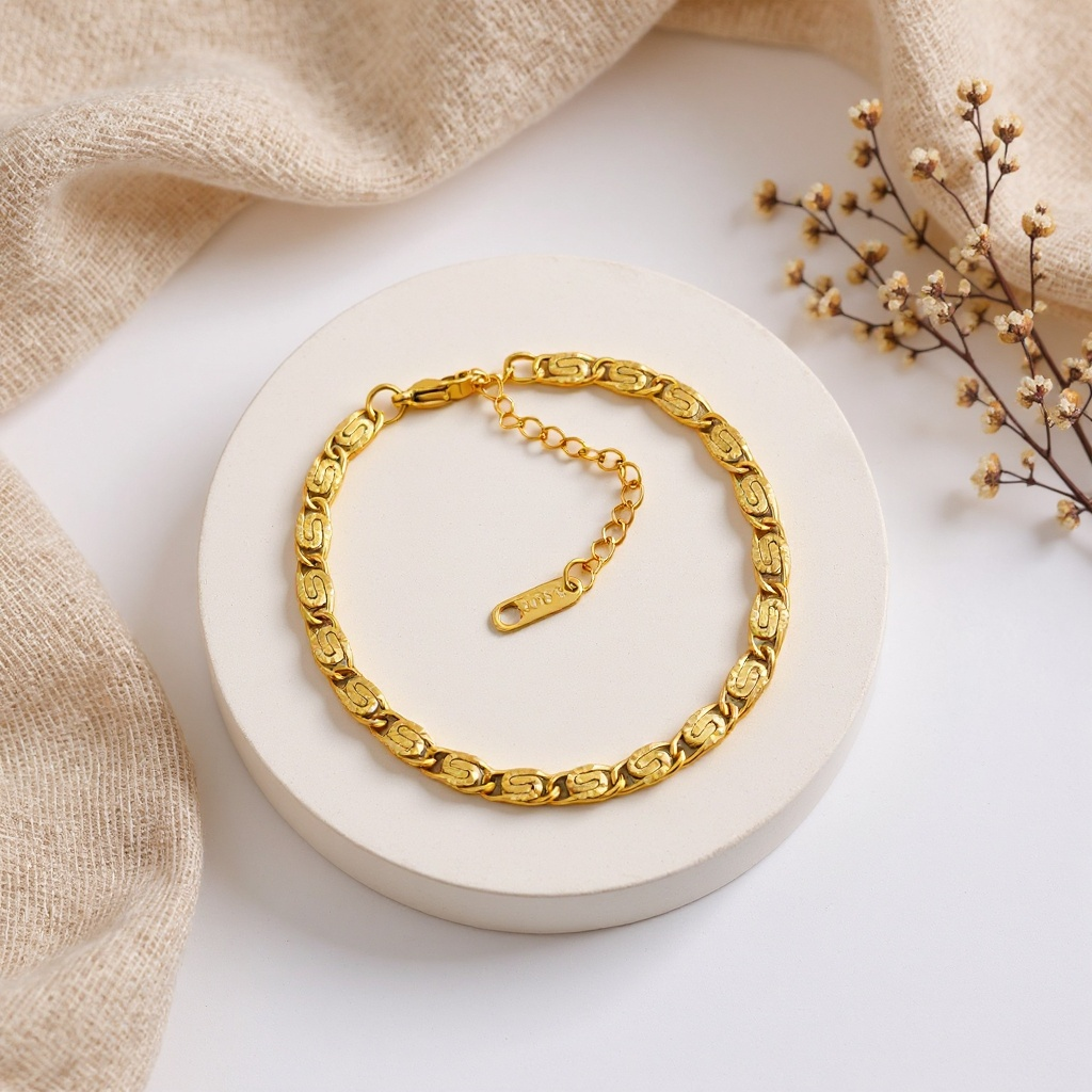 Gold chain bracelet on a white surface with beige fabric and dried flowers.
