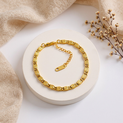 Gold chain bracelet on a white surface with beige fabric and dried flowers.