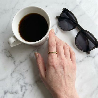 White mug with dark liquid, sunglasses, and a hand with a gold minimalist ring on a marble surface