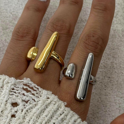 Hand wearing two chunky rings, one gold and one silver, on a neutral background.