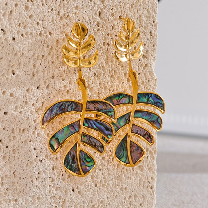 Gold leaf-shaped earrings with abalone shell inlay on a textured beige surface.