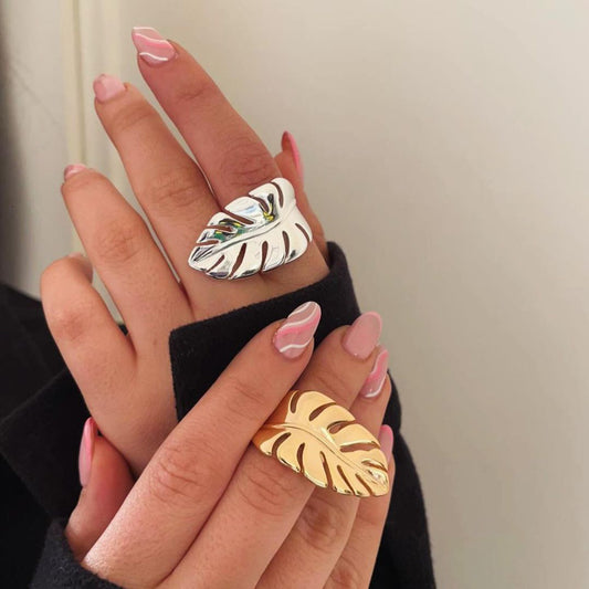 Two hands wearing large leaf-shaped rings, one gold and one silver, against a neutral background.