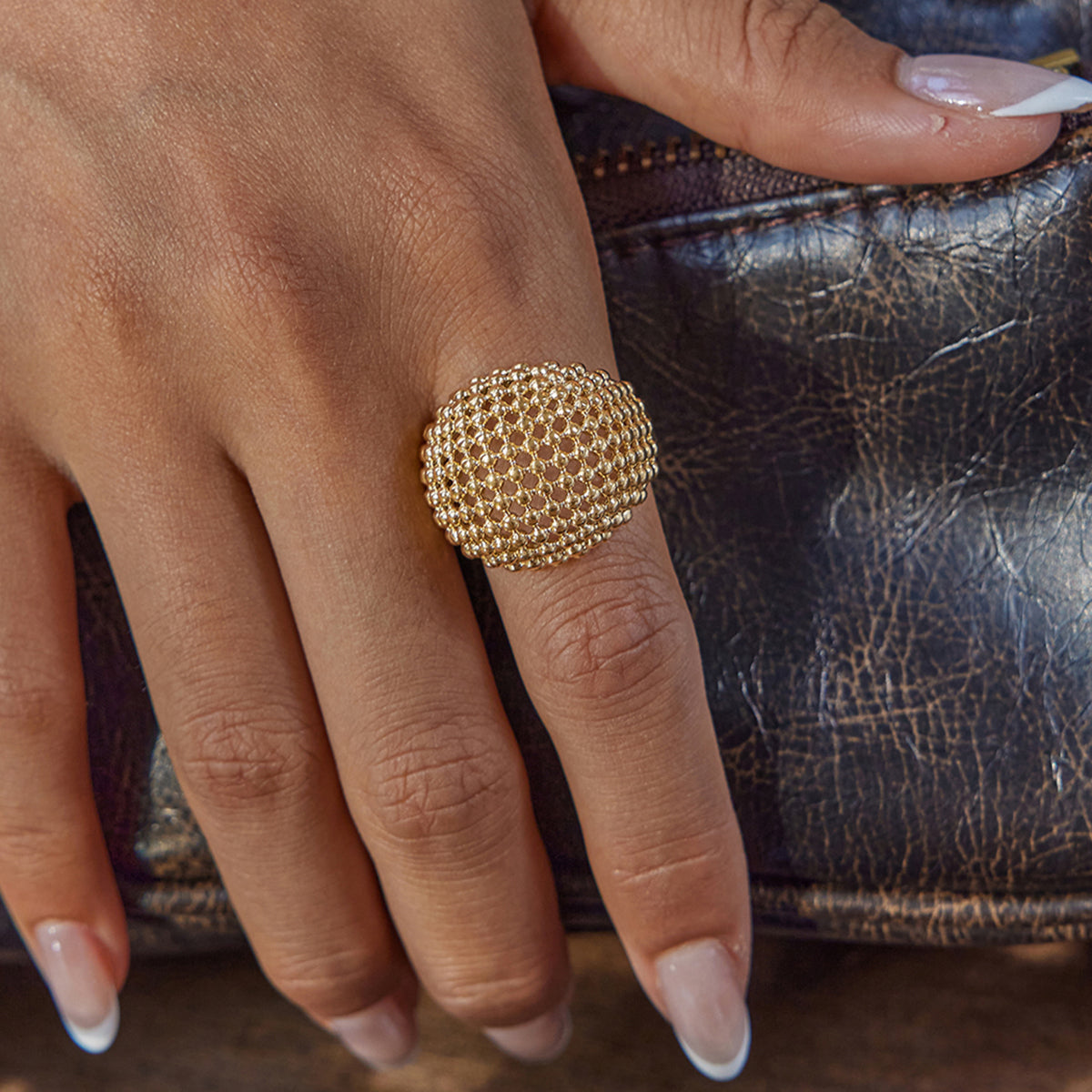 Gold statement ring on a finger with a textured design, held against a textured surface.