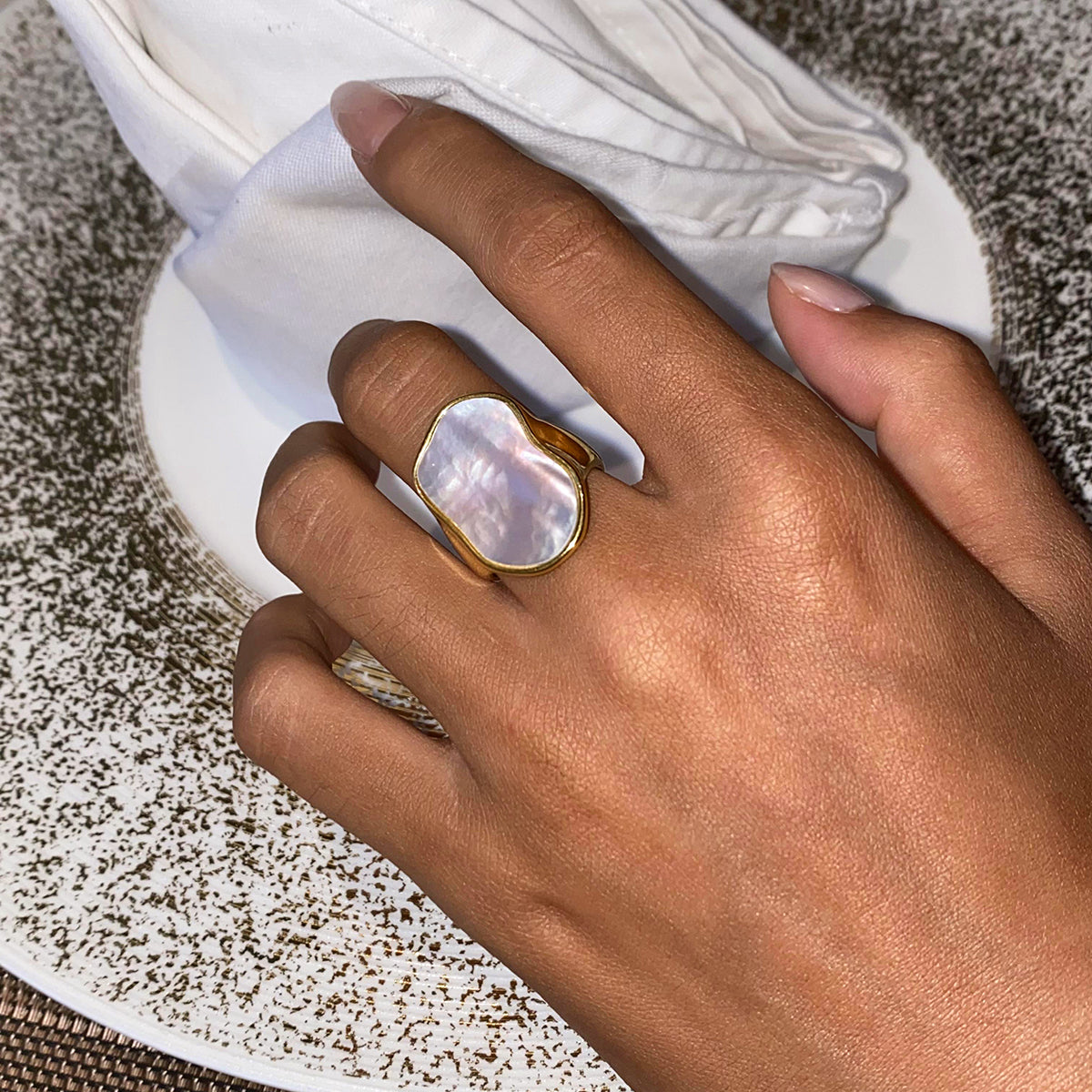Hand wearing a gold chunky ring with a white stone on a textured surface