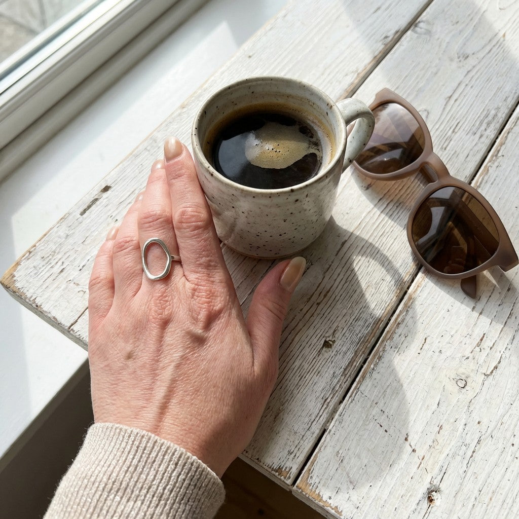 Hand wearing waterproof silver ring holding a coffee mug on a wooden surface with sunglasses nearby