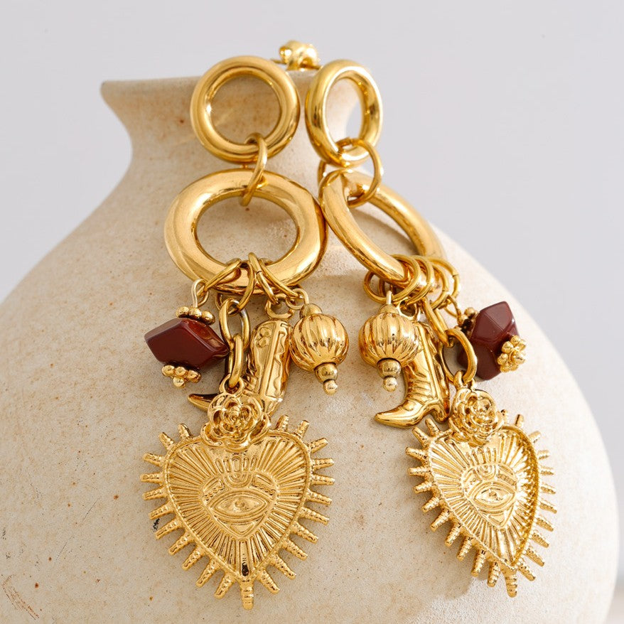 Gold earrings with heart-shaped charms and red stones on a beige stone surface