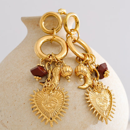Gold earrings with heart-shaped charms and red stones on a beige stone surface