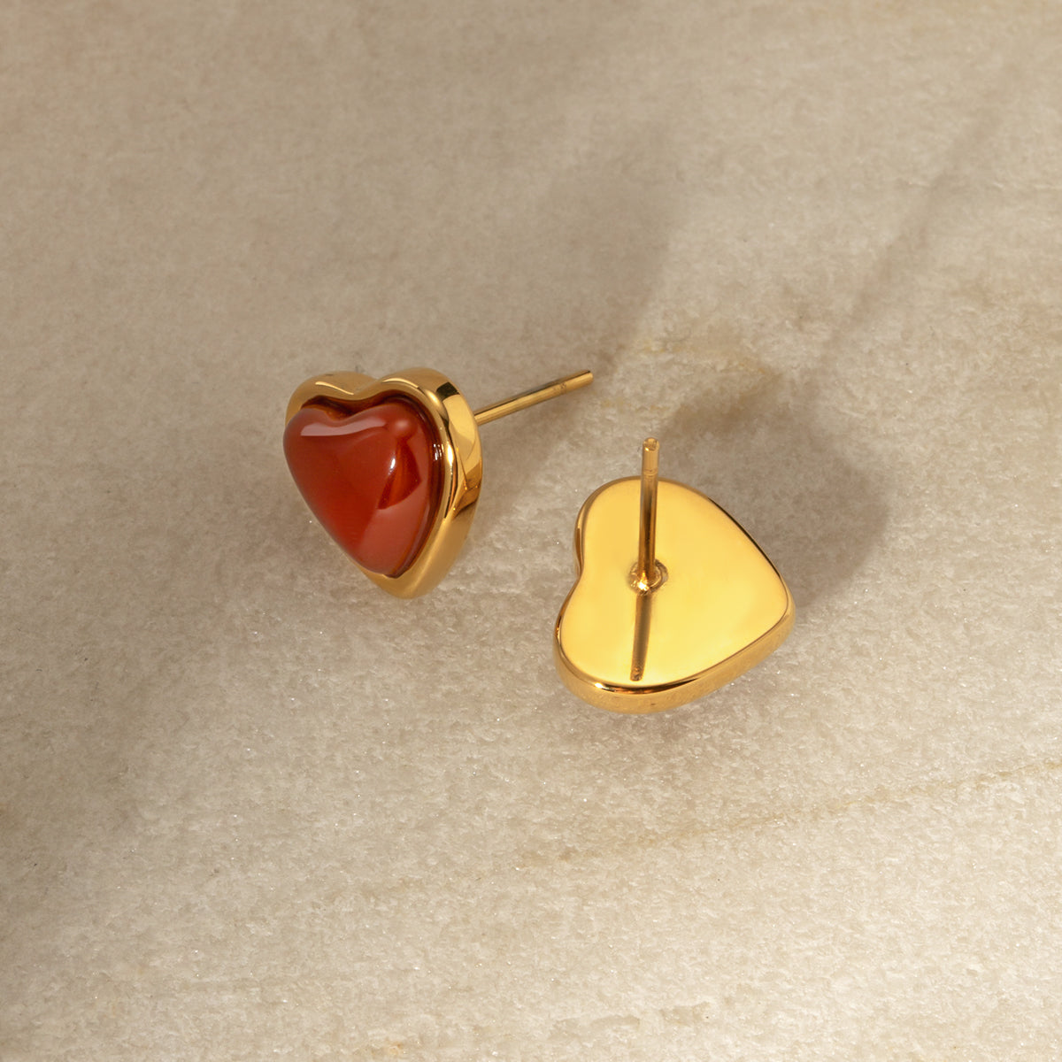 Heart-shaped stainless steel earrings with gold frame and red stone on beige background