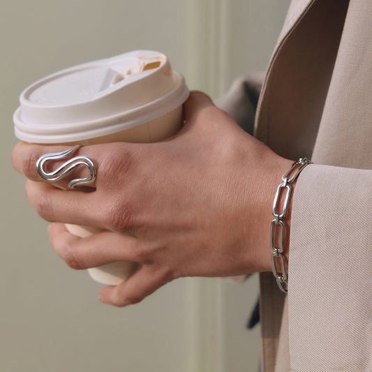 Hand holding a coffee cup with silver ring and bracelet on a neutral background