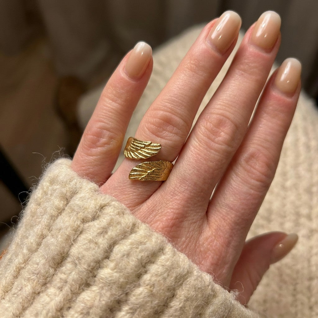 Hand wearing gold leaf-shaped ring with beige nail polish against a blurred background.